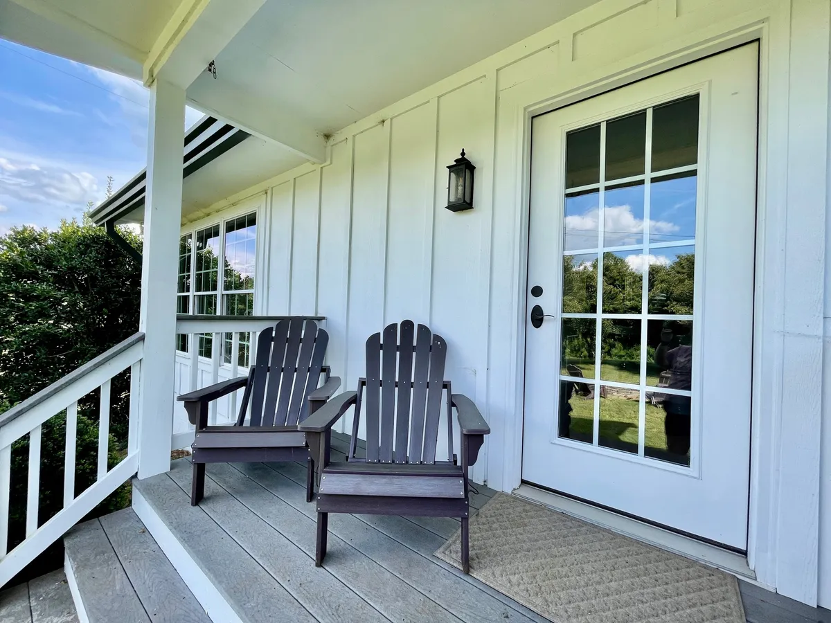 The image features two Adirondack chairs on a welcoming front porch, bathed in soft morning light. A decorative lantern and the glass-paneled front door add charm, while the chairs are perfectly positioned to enjoy a peaceful sunrise or sunset. The mood is intimate and inviting, ideal for couples seeking relaxation.