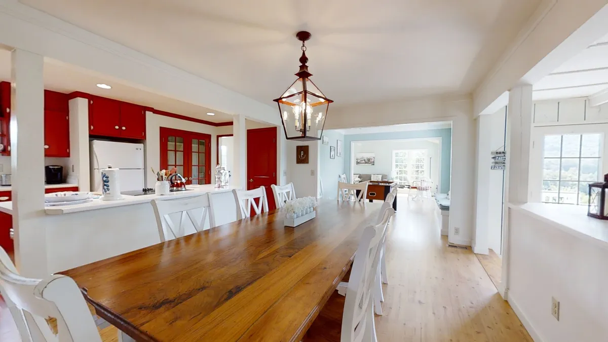 A long wooden dining table set for eight stretches beneath a rustic lantern-style light fixture. The adjacent kitchen area features the same red cabinetry, tying the two spaces together. White dining chairs and farmhouse accents complete the room’s warm, communal vibe.