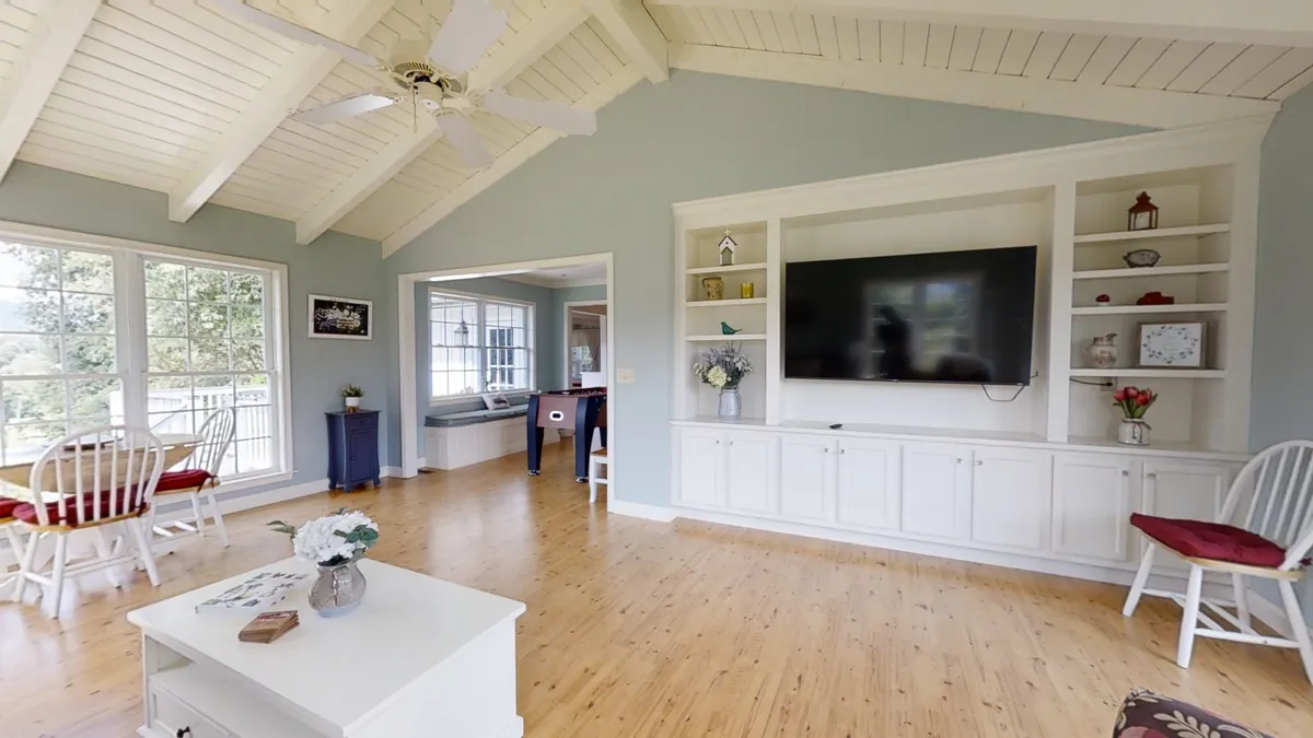 A bright, airy living space featuring large windows, a TV unit, a dining area, and comfortable seating. Sunlight streams in, and the open layout suggests easy flow between gathering, dining, and relaxing.
