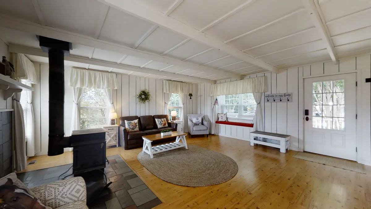 This image showcases a living area with a wood-burning stove as a focal point, comfortable plush seating clustered around it, and large windows letting in abundant natural light. The ambiance is cozy and welcoming, ideal for relaxing, socializing, or enjoying a board game with family and friends.