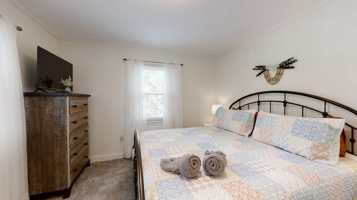 A cozy bedroom featuring a comfortable bed with soft linens, a dresser for storage, a window allowing natural light, and neatly arranged towels. The overall mood is warm and restful, perfect for unwinding after a long day.