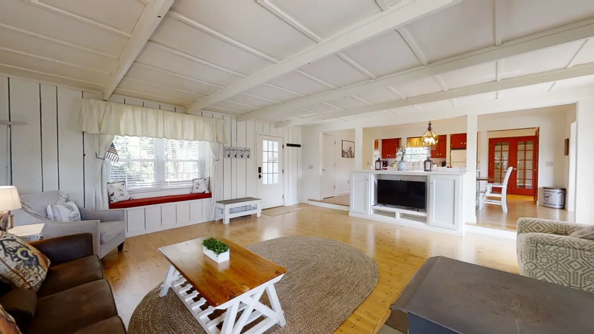A comfortable living space with a large window seat, a cozy sofa, and a wooden coffee table, set against a backdrop of a kitchen area. The room is filled with natural light, offering a welcoming spot for relaxation and casual gatherings.
