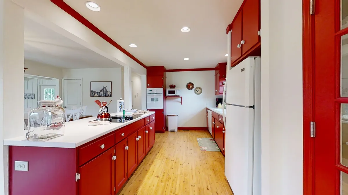 The kitchen is shown from a different perspective, emphasizing the continuity of red cabinetry, white countertops, and wooden flooring. Kitchen appliances and functional workspace speak to modern convenience, while the inviting color scheme maintains a cheerful ambiance.